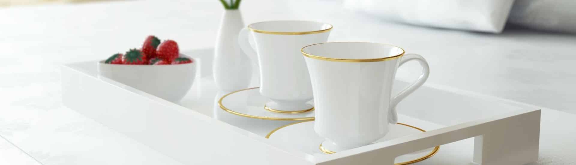 White tray on a bed with gold rimmed teacups and saucers, yellow flower in a vase and a bowl of strawberries