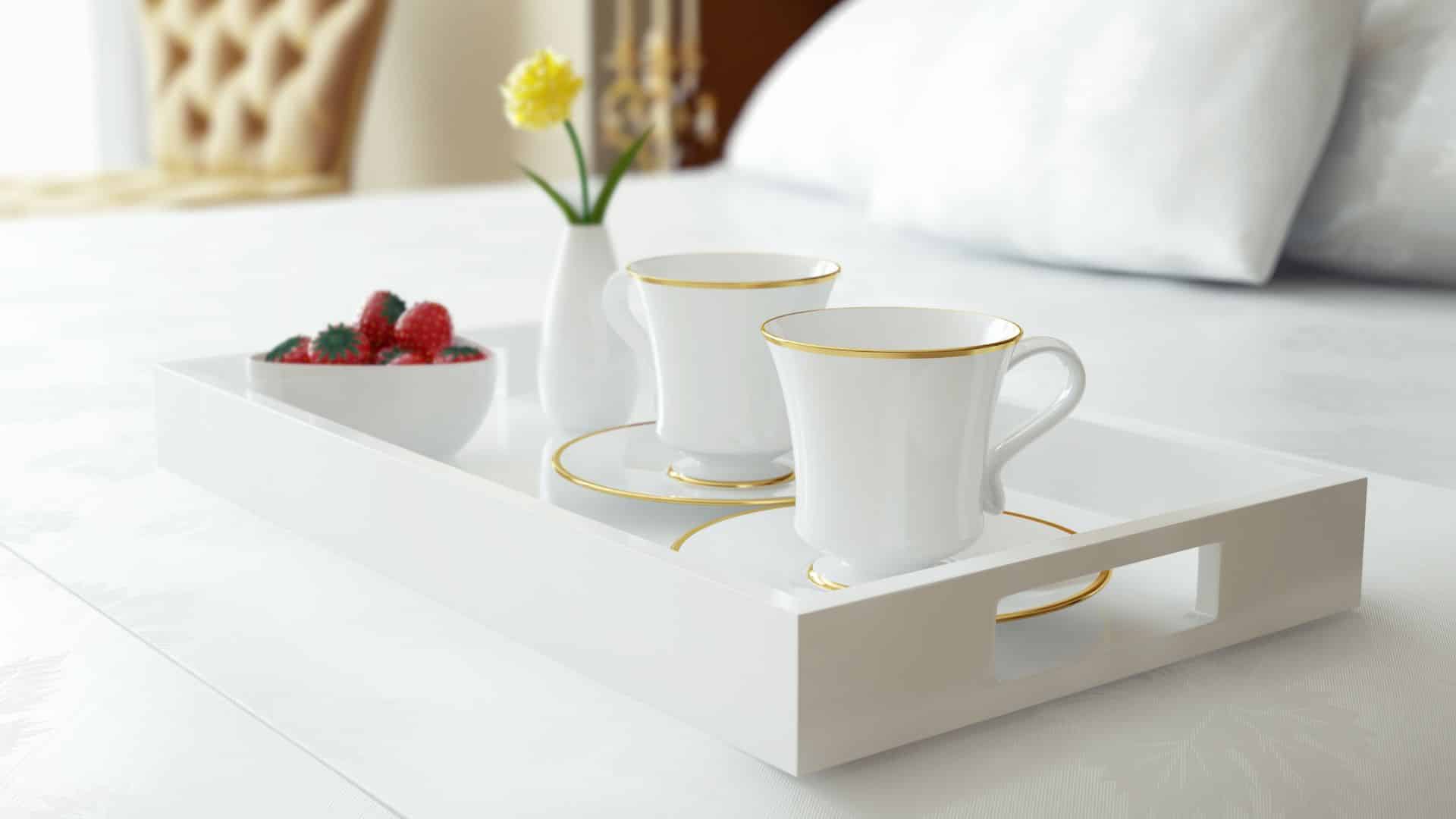 White tray on a bed with gold rimmed teacups and saucers, yellow flower in a vase and a bowl of strawberries