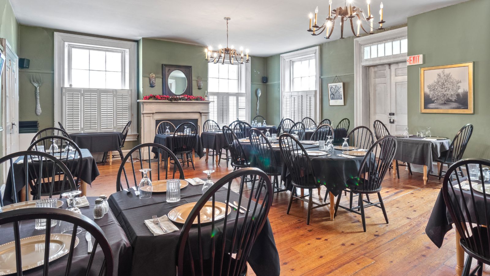 Large dining room with bright windows, hardwood floors and several tables set with gold plates and black tablecloths