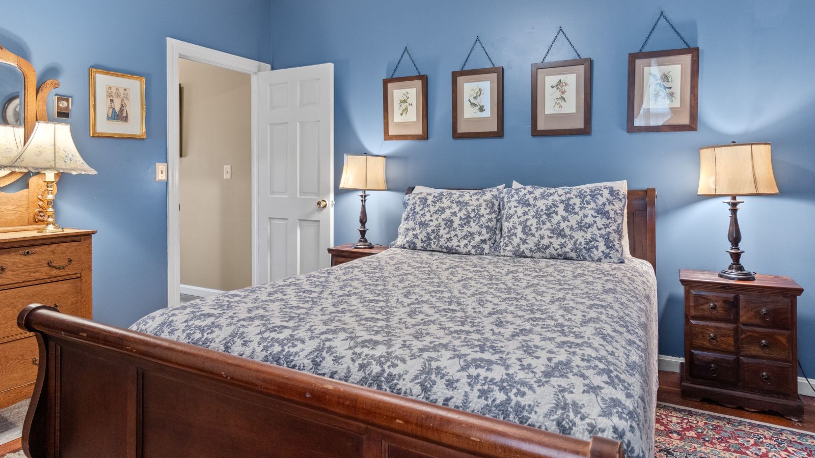 Bedroom with queen sleight bed, blue walls, colorful rug, side tables with lamps and artwork