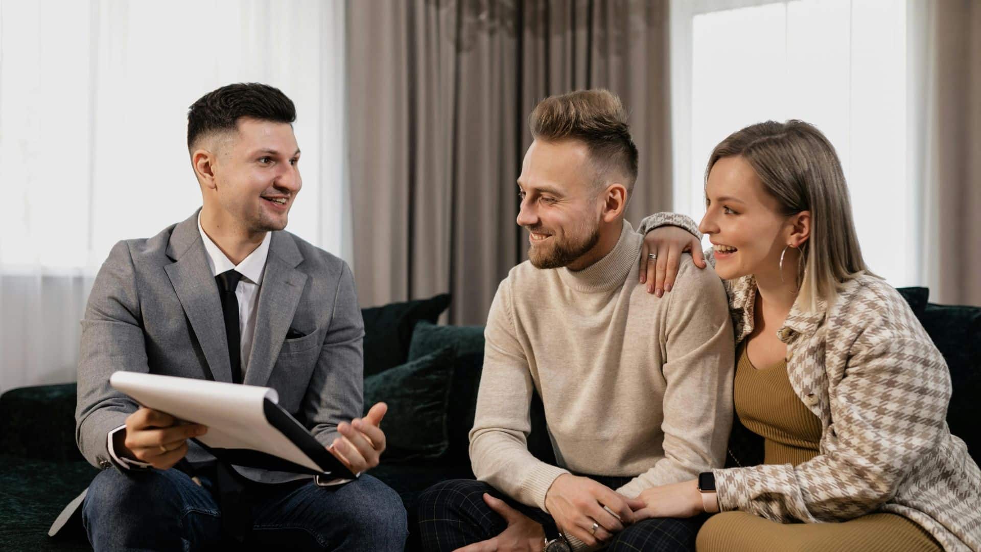 A man in a grey suit talking to a couple on a couch by windows with curtains