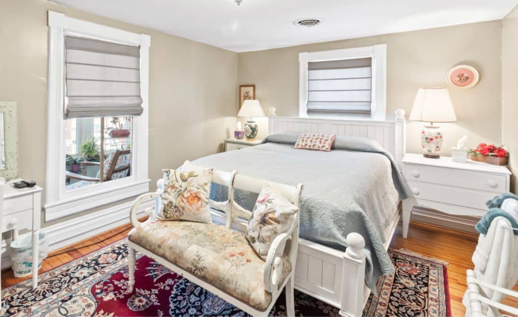 Elegant bedroom with queen bed, white furniture, oriental rig and windows with roman shades