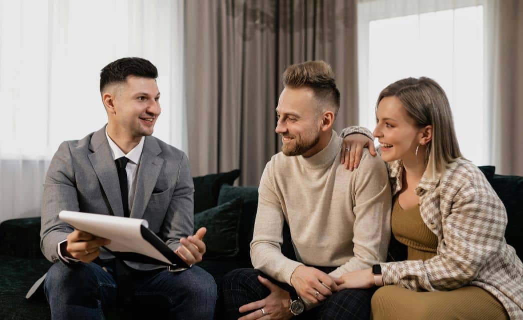 A man in a grey suit talking to a couple on a couch by windows with curtains