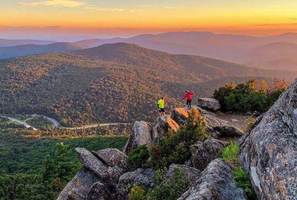 Two hikers perched on large rocks at the edge of a cliff overlooking an expansive tree covered mountain range